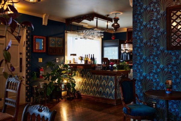 A wooden bar with a teal and turquoise, peacock-feather-like pattern is lined with bottles of wine. It looks onto a small dining room with teal and royal blue notes, from a regal-looking chair to a wallpaper with bouquets of midnight blue flowers.