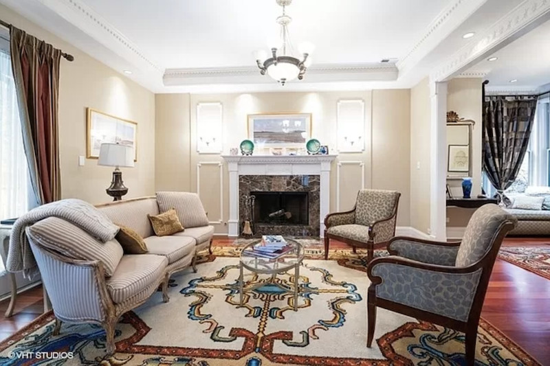 An ornate living room with a pattern carpet, footed couch and chairs. and a stone fireplace. 