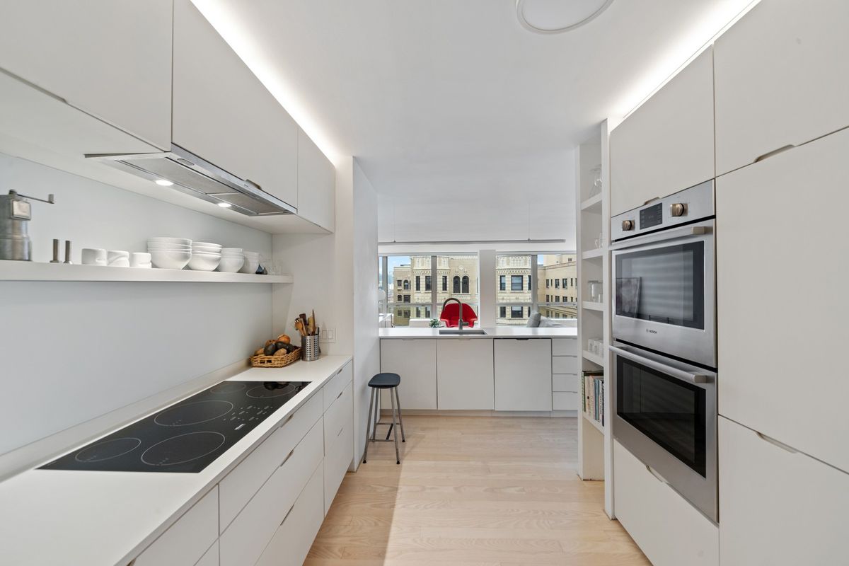A gallery style-kitchen with modern white cabinets, integrated metal appliances, and hardwood floors. 