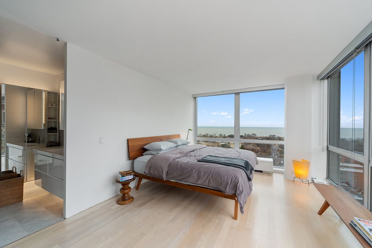 A corner bedroom with a contemporary wood framed bed and floor windows overlooking a park and large body of water. 