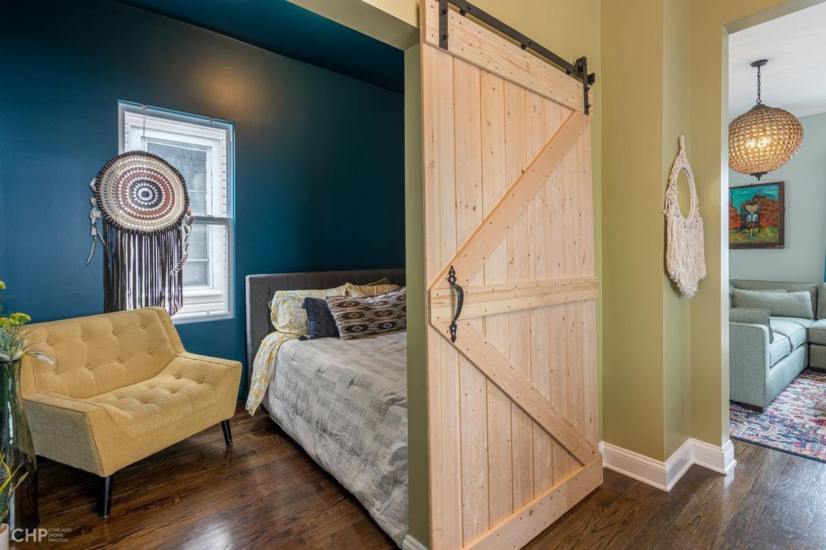 A sliding barn door reveals a bedroom with a blue wall and a cream colored chair. 