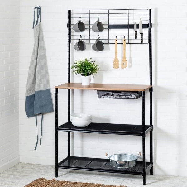 A kitchen rack with two open storage shelves, a small drawer for dishtowels, a wooden counter shelf, and a grid rack for holding knives, serving utensils, and mugs