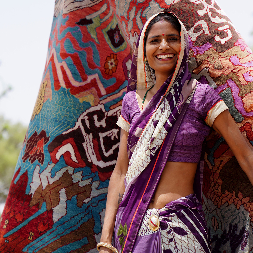 Indian woman smiling and wearing purple, standing in front of a multicolored hand-knotted rug that is available at Cyrus Artisan Rugs