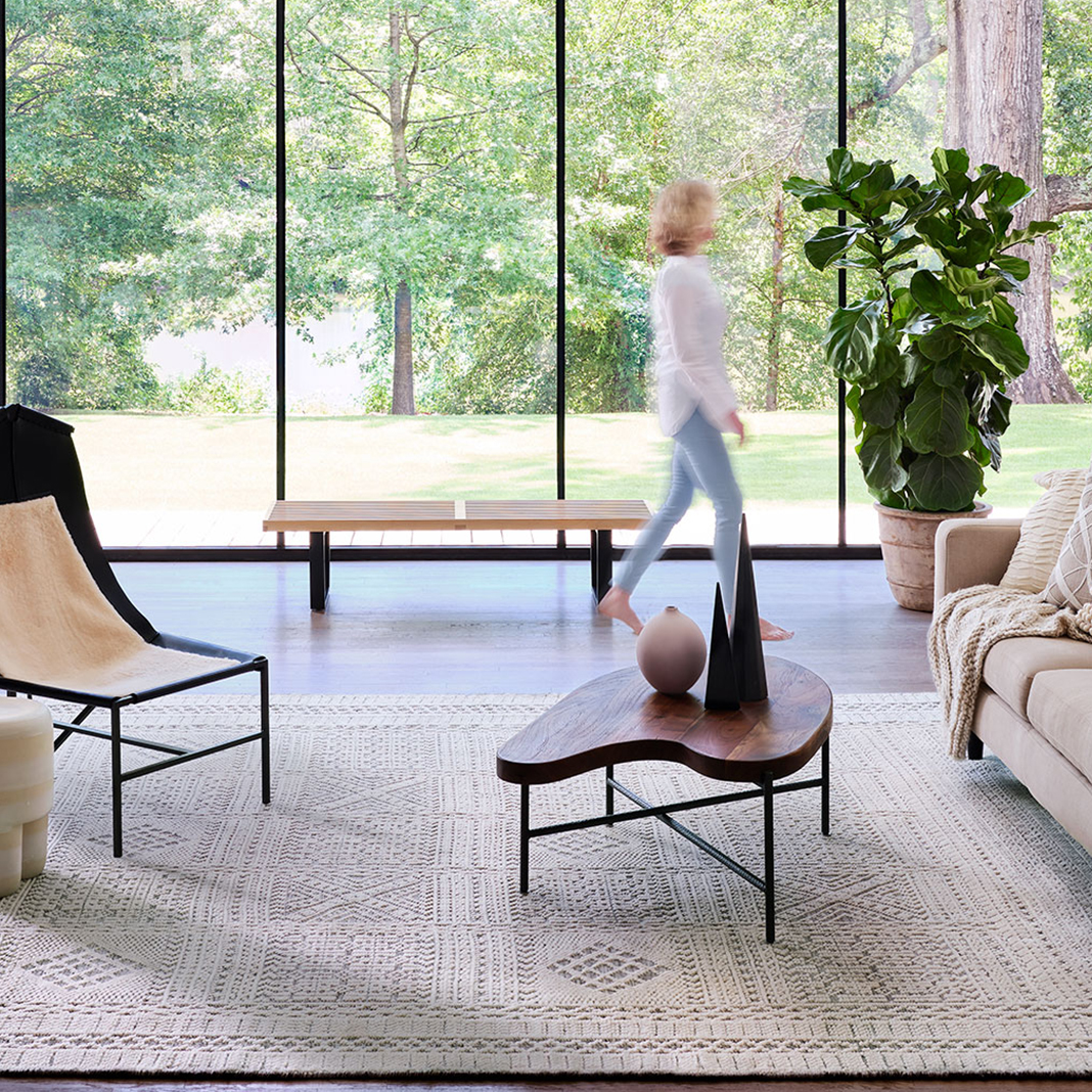 Woman walking through bright living room with light colored couch and chair and a tan wool rug from Cyrus Artisan Rugs